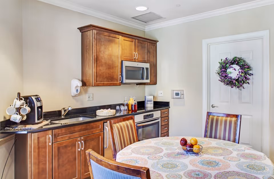 A small kitchen area with wooden cabinets, a microwave, an oven, and a sink. On the countertop, there is a coffee maker with hanging mugs, a basket with glasses, and bottles of juice. In front of the kitchen is a round table covered with a colorful patterned tablecloth and surrounded by wooden chairs with striped cushions. A door with a decorative purple and green wreath is visible in the background.