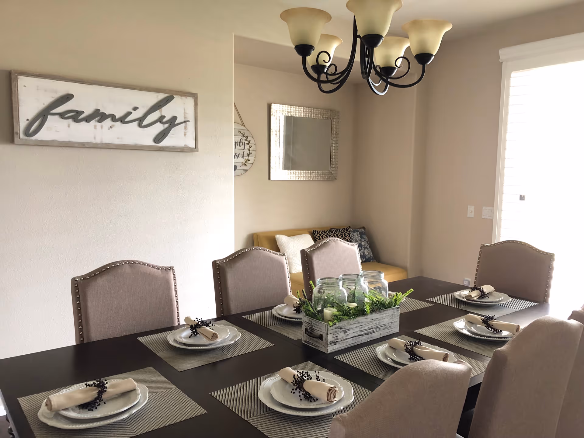 A dining room with a dark wooden table set for six people. Each place setting has a white plate, a rolled beige napkin with a black decorative ring, and a striped placemat. In the center of the table is a rustic wooden box with three glass jars and green foliage. Beige upholstered chairs surround the table. On the wall, there is a decorative sign that says 'family' and a large mirror. A chandelier with five frosted glass shades hangs above the table. Light enters the room through a window with white blinds.