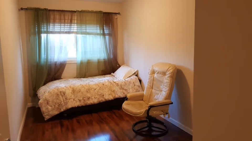 A small bedroom with a single bed covered in a floral comforter positioned against the wall under a window with sheer brown curtains. A beige leather swivel chair is placed near the bed on a wooden floor.