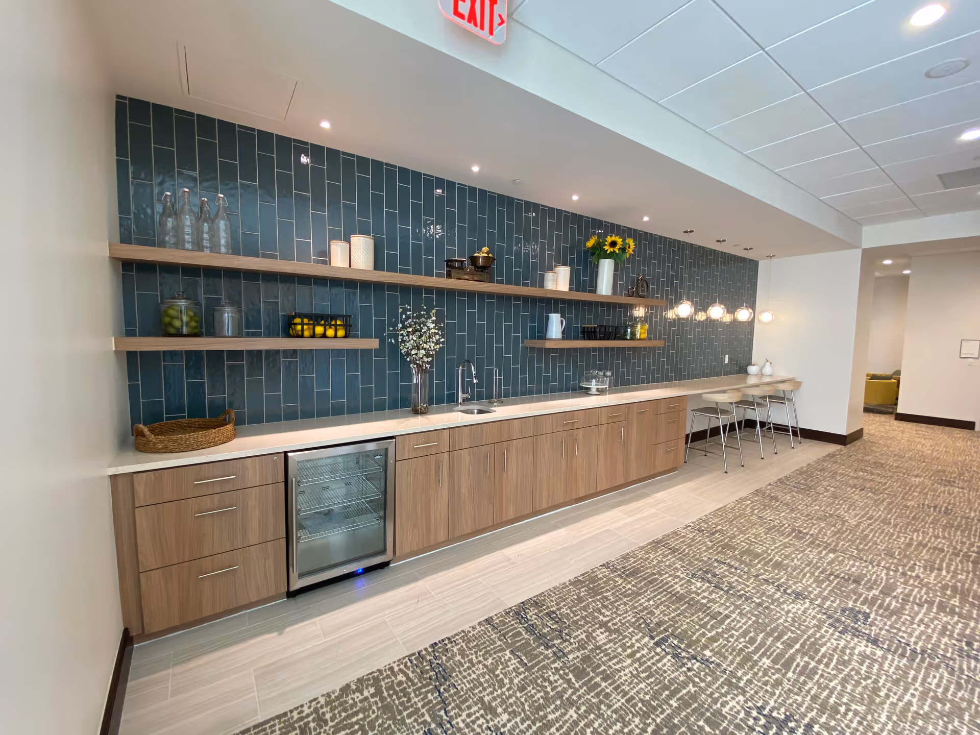 A modern kitchenette area with wooden cabinets, a small refrigerator, and a sink. Above the counter are two wooden floating shelves holding decorative items such as glass bottles, jars with lemons and olives, a vase with flowers, and a pitcher. The backsplash is made of vertical dark blue tiles. To the right, there is a counter with three bar stools and hanging globe pendant lights. The floor is a combination of light tiles and patterned carpet, and an exit sign is visible on the ceiling.