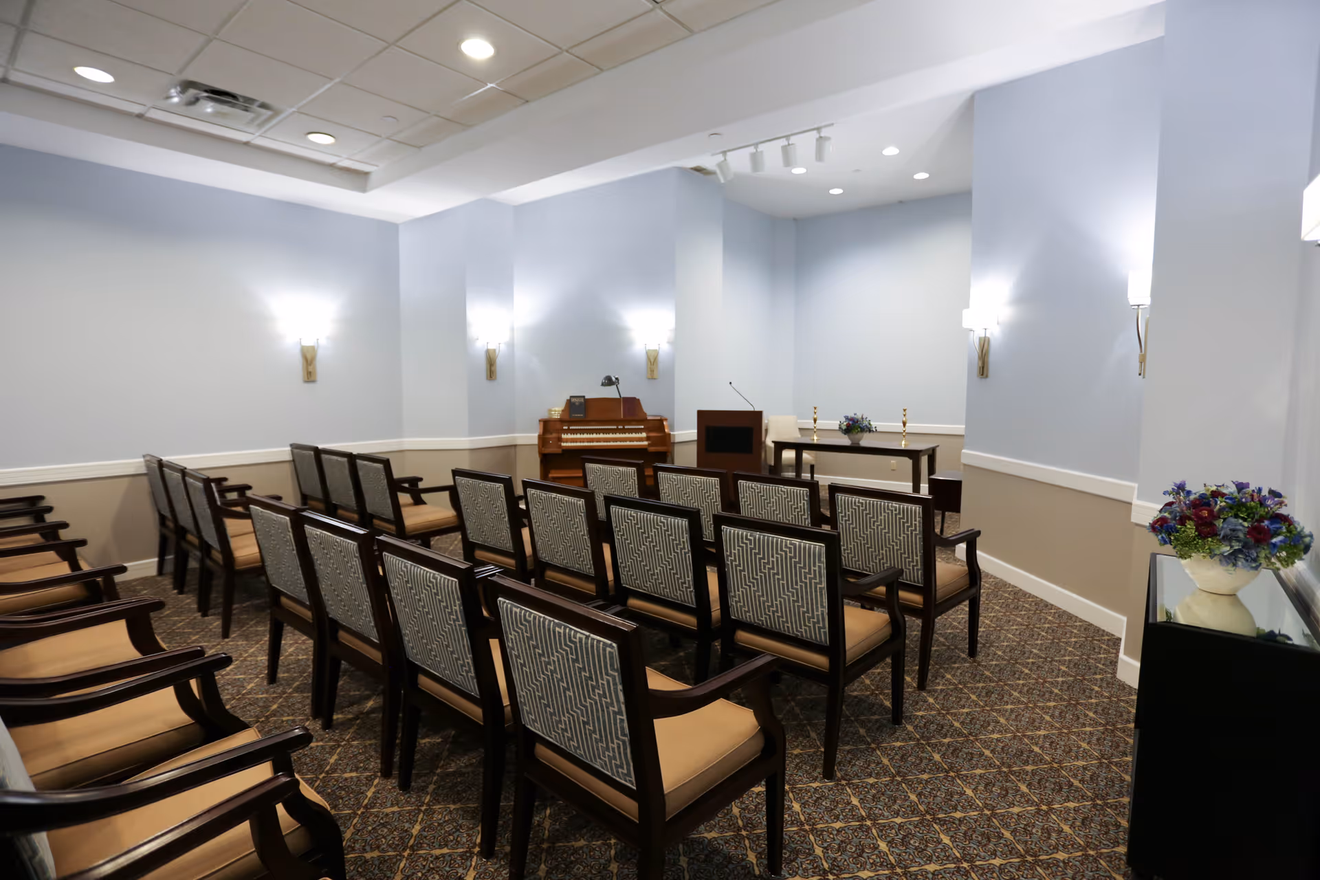 A small chapel or meeting room with rows of wooden chairs with patterned backs and beige cushions facing a wooden podium and a piano. The walls are painted light blue with beige wainscoting, and there are wall-mounted lights and a floral arrangement on a side table.