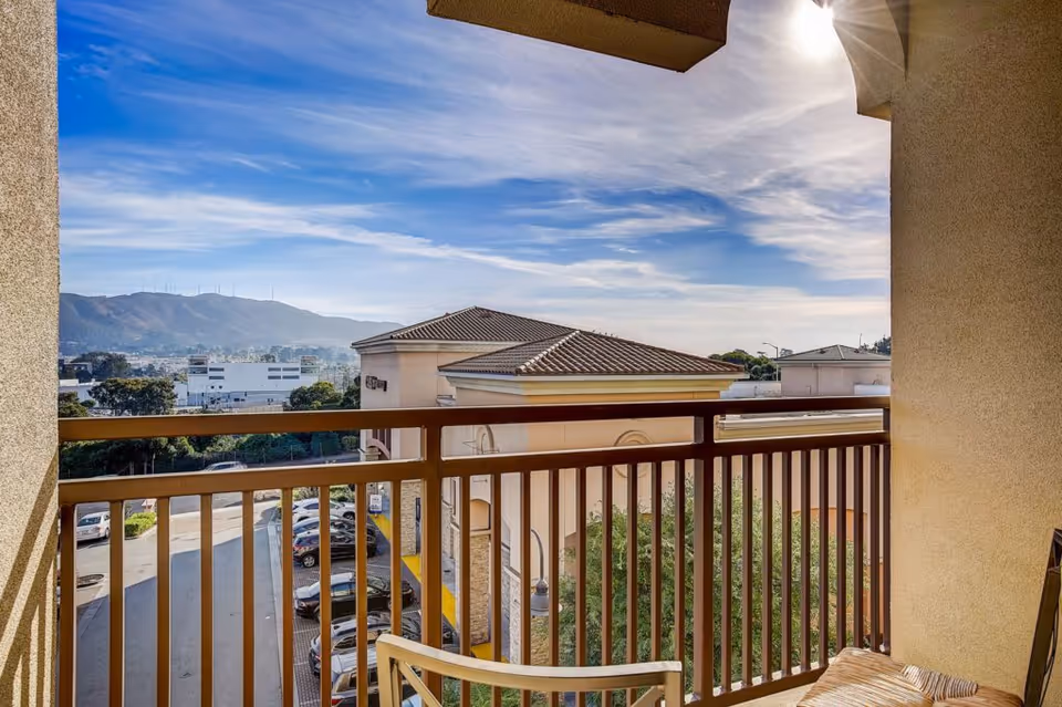 View from a balcony showing a railing, a chair, nearby buildings with tiled roofs, parked cars, trees, and hills in the distance under a partly cloudy blue sky.