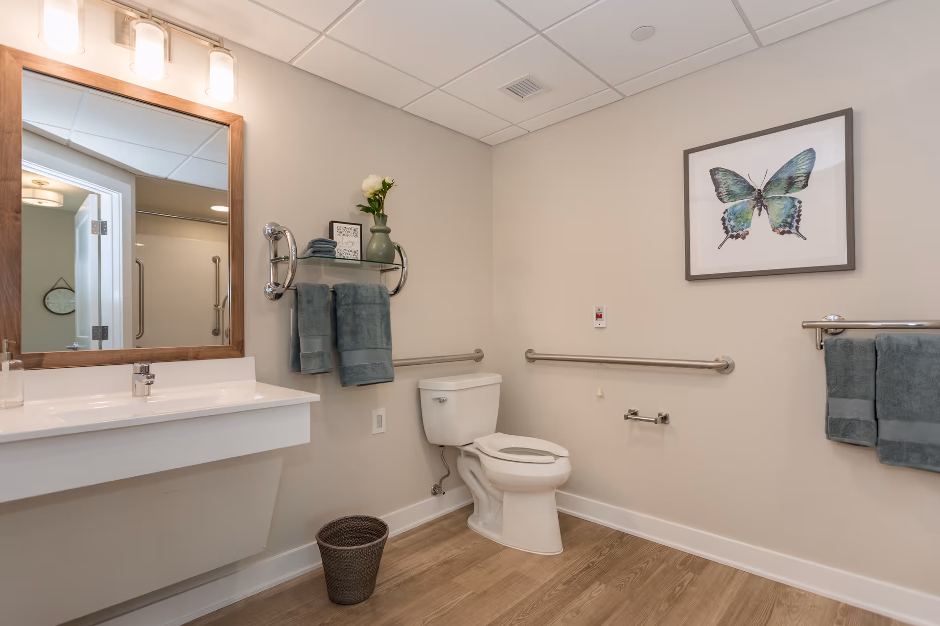 A clean and modern bathroom with a white toilet, a white sink with a wooden framed mirror above it, and a shelf holding folded towels and a vase with flowers. There are grab bars on the walls for accessibility, a framed butterfly artwork on the wall, and a small wastebasket on the wooden floor.