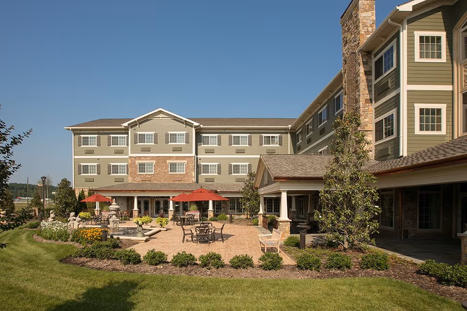 Exterior view of a multi-story senior living building with a landscaped courtyard, patio tables and red umbrellas, and a fountain.