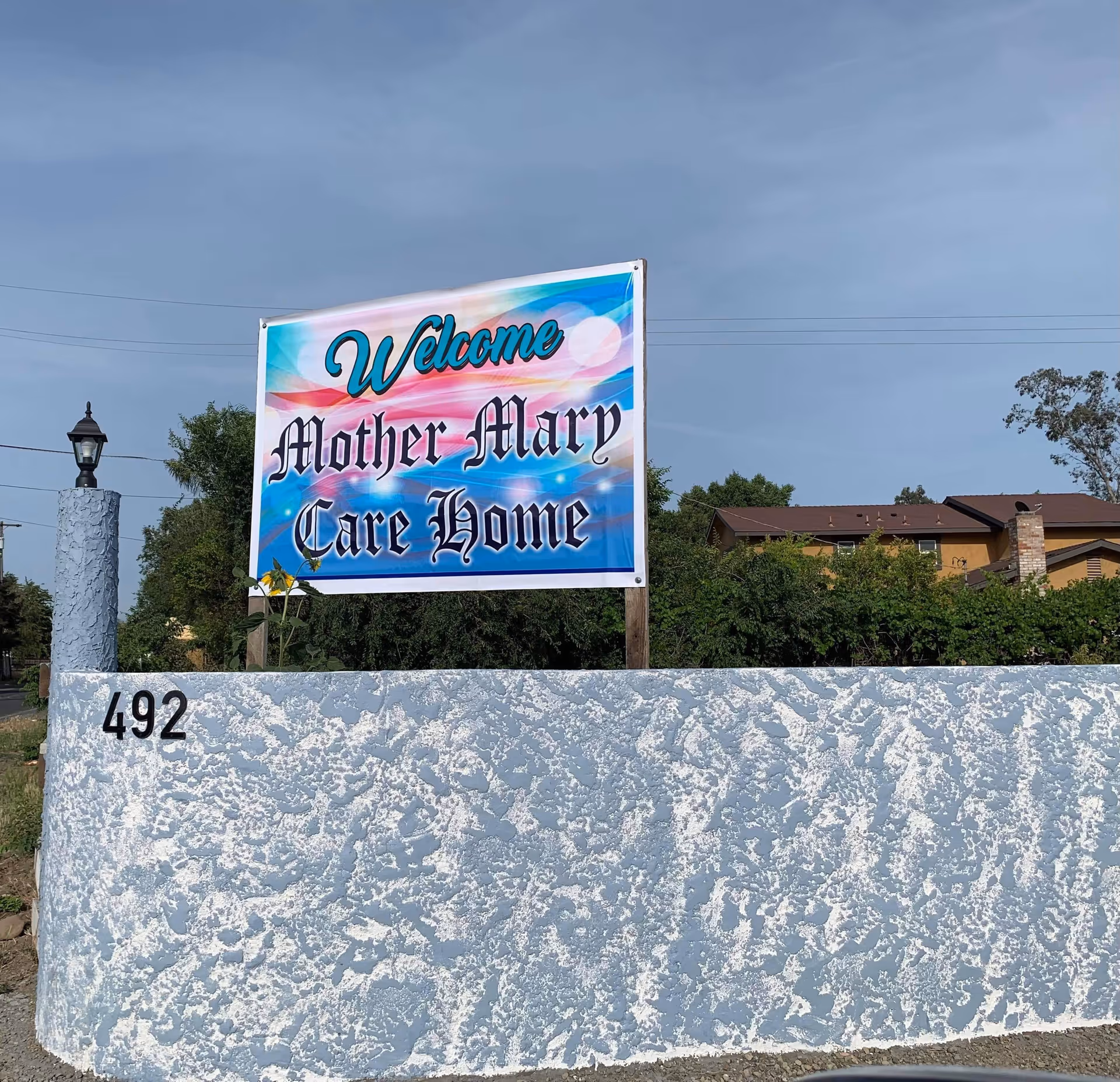 A colorful roadside sign reading "Welcome Mother Mary Care Home" mounted above a textured low wall marked with the number 492 and a nearby lamp post.