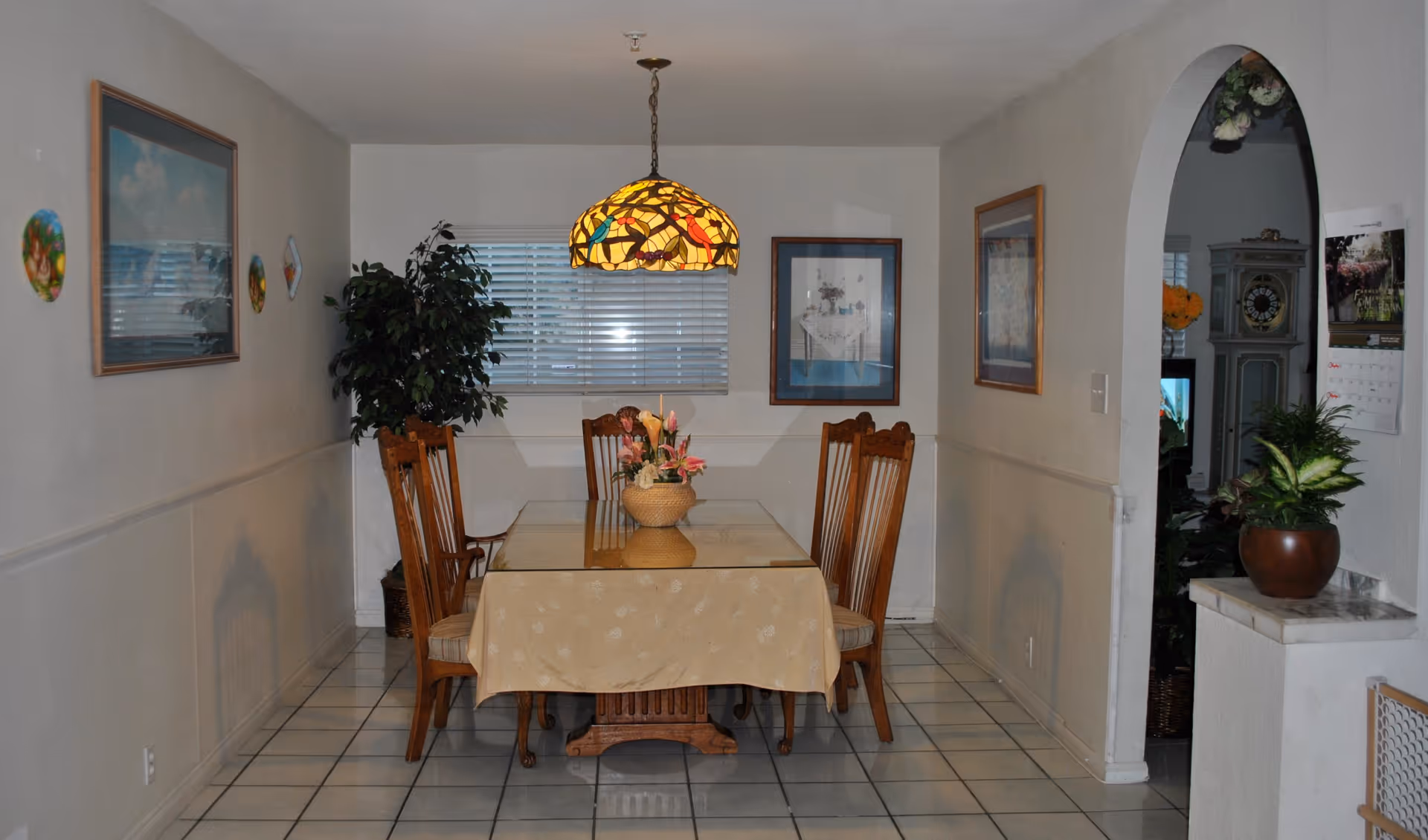 A dining room with a rectangular table covered by a beige tablecloth and a glass top, surrounded by six wooden chairs. A stained glass pendant light hangs above the table. The room has white tiled flooring and white walls adorned with framed pictures and decorative plates. A potted plant is placed in the corner near a window with blinds, and an arched doorway leads to another room with a grandfather clock and more plants.