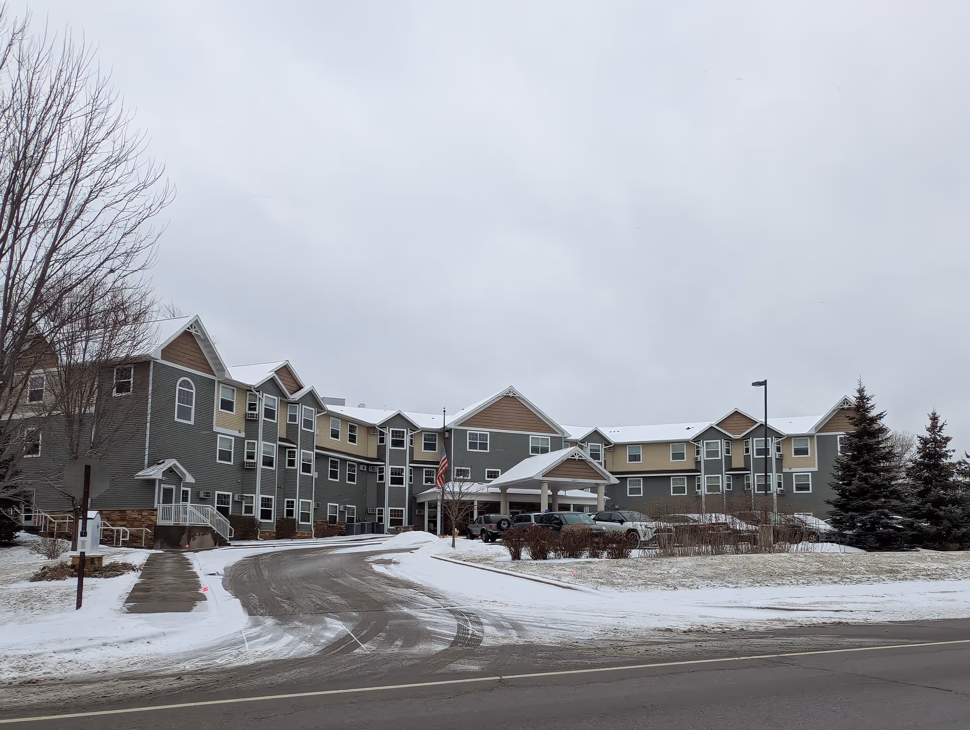 Three-story apartment-style senior living building with a covered main entrance, parked cars, and light snow on the ground.