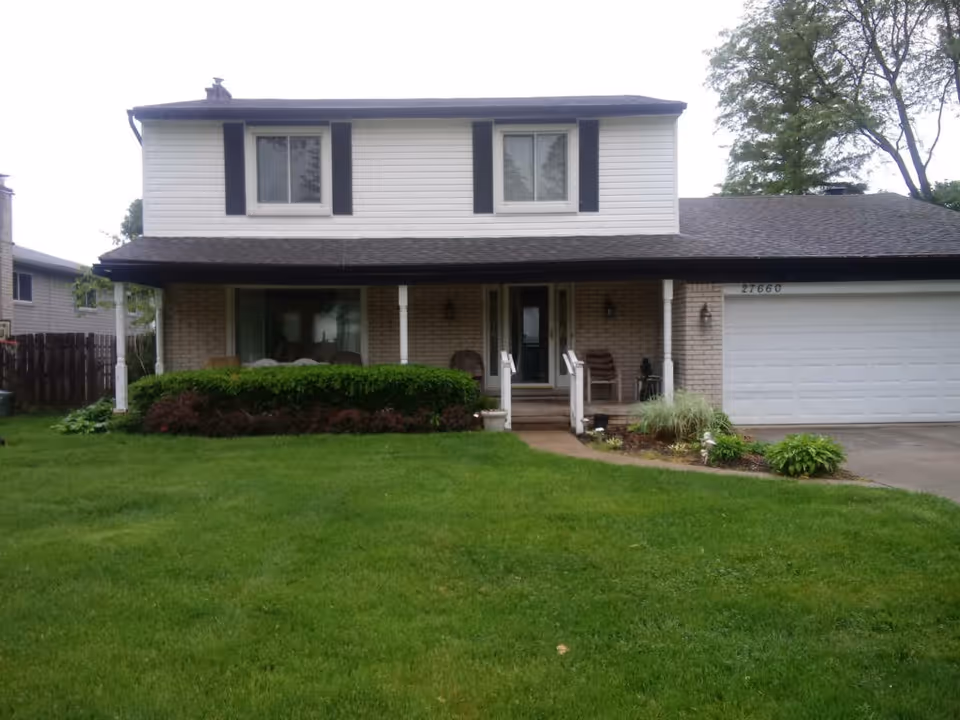 Front exterior view of a two-story house with white siding and brick facade, black shutters on the upper windows, a covered porch with chairs, a well-maintained green lawn, and a driveway leading to a white garage door.