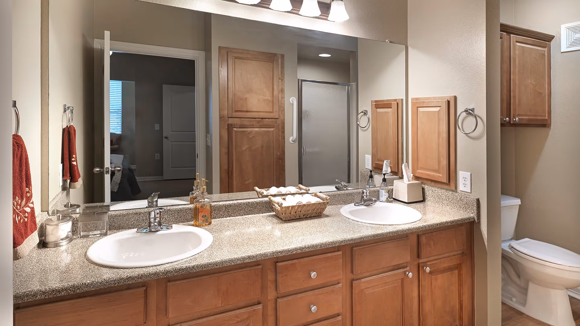 A bathroom with a double sink vanity featuring a granite countertop, wooden cabinets, and a large mirror above. There are red hand towels hanging on towel rings on the left wall. A basket with rolled white towels is placed between the sinks. In the background, a toilet and a shower with a glass door are visible.