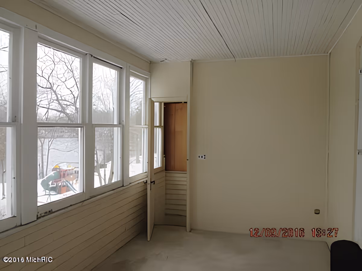 Empty sunroom or enclosed porch with large windows on the left side showing a snowy outdoor scene with trees and a playground. The room has beige walls, a white paneled ceiling, and a small door slightly ajar on the far wall.