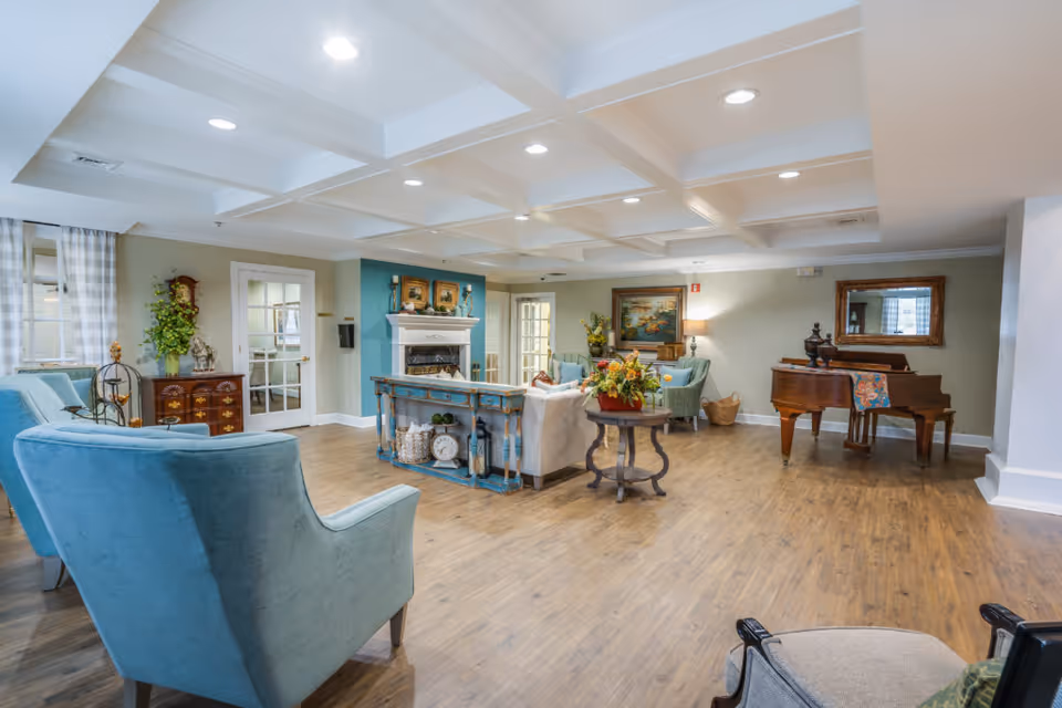 Bright, spacious common living room with teal armchairs, a fireplace, a grand piano, and decorative furnishings.