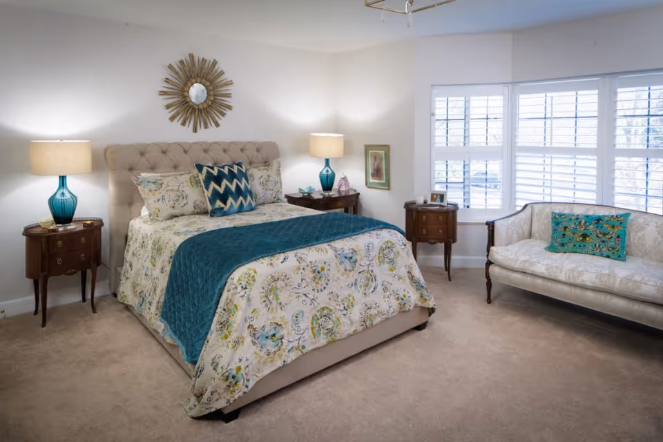 A well-lit bedroom with a beige tufted bed adorned with floral bedding and a teal throw blanket. Two wooden nightstands with teal glass lamps flank the bed. A decorative sunburst mirror hangs above the bed. To the right, there is a cream-colored upholstered loveseat with a teal decorative pillow in front of large windows with white shutters.