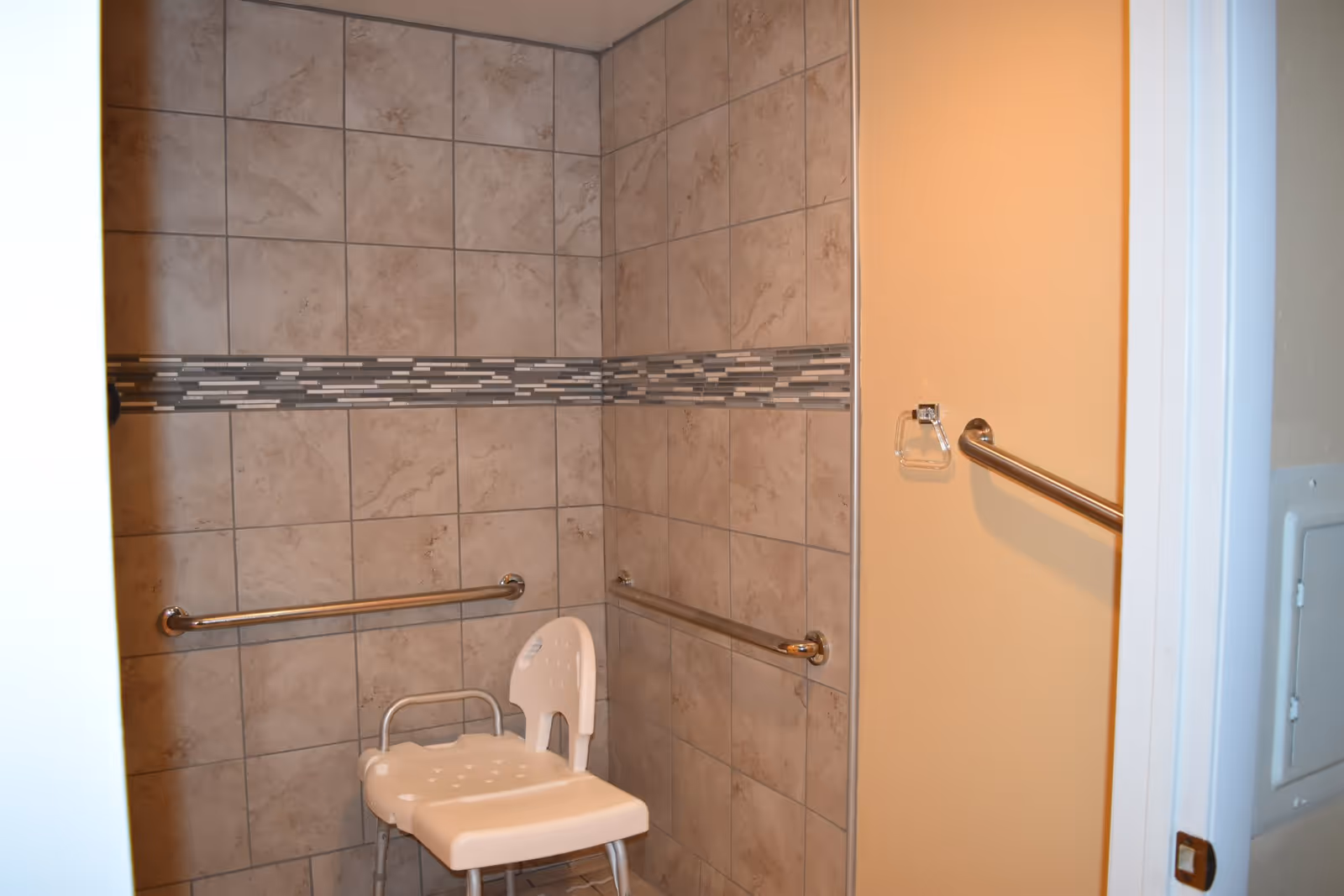 A tiled shower area with beige and brown tiles, featuring a horizontal decorative strip of smaller rectangular tiles. There are two metal grab bars mounted on the walls and a white plastic shower chair with a backrest inside the shower. A towel ring is mounted on the adjacent wall outside the shower.