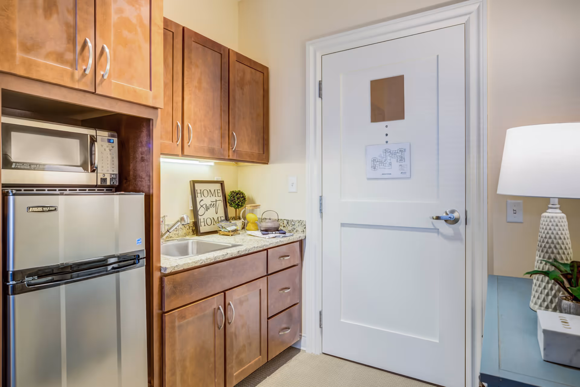 Small kitchenette area with wooden cabinets, a stainless steel mini refrigerator, a microwave, a sink, and a countertop with decorative items including a framed 'Home Sweet Home' sign, a small plant, and a lamp on a side table next to a white door.