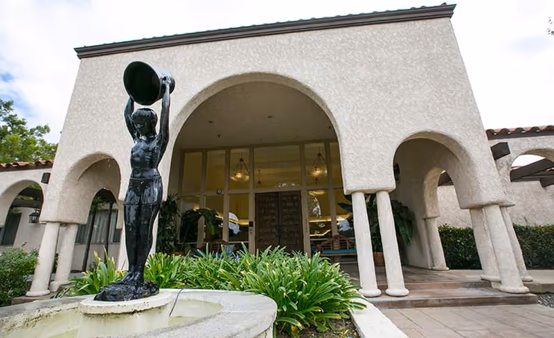 Exterior view of Silverado Calabasas Memory Care Community featuring a beige stucco building with arched entryways and columns. In front of the building is a black statue of a person holding a large bowl above their head, positioned in the center of a small fountain surrounded by green plants.