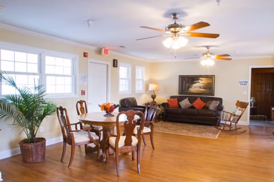 A bright and spacious living room area with wooden flooring, featuring a round wooden dining table with four chairs, a large potted plant near the window, a dark leather sofa with colorful cushions, a rocking chair, a floor lamp, and ceiling fans with lights. There is a painting on the wall and multiple windows letting in natural light.
