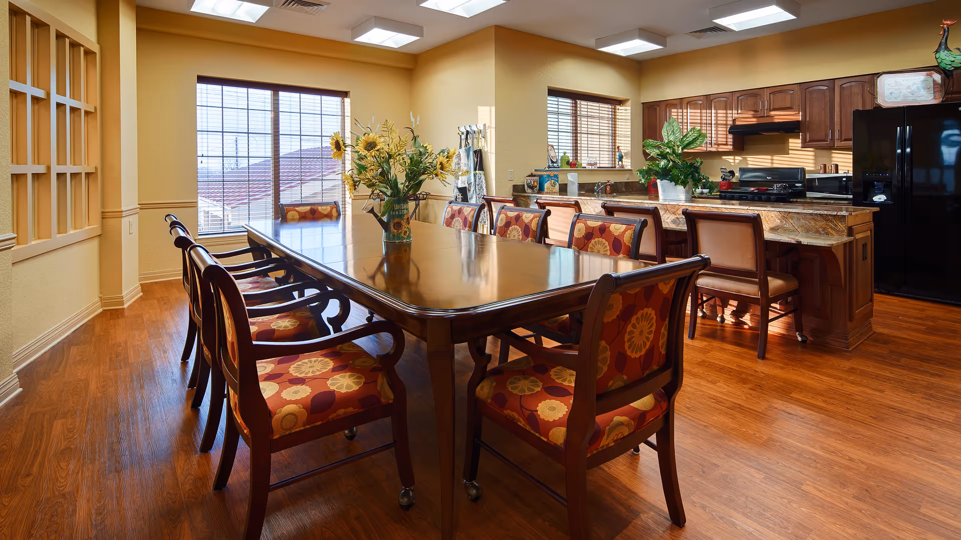 A bright dining area with a long wooden table surrounded by chairs with floral patterned cushions. The room features large windows with blinds allowing natural light to fill the space. Adjacent to the dining area is a kitchen with wooden cabinets, a black refrigerator, a stove, and a marble countertop with bar stools. There are decorative plants and a vase with sunflowers on the table.