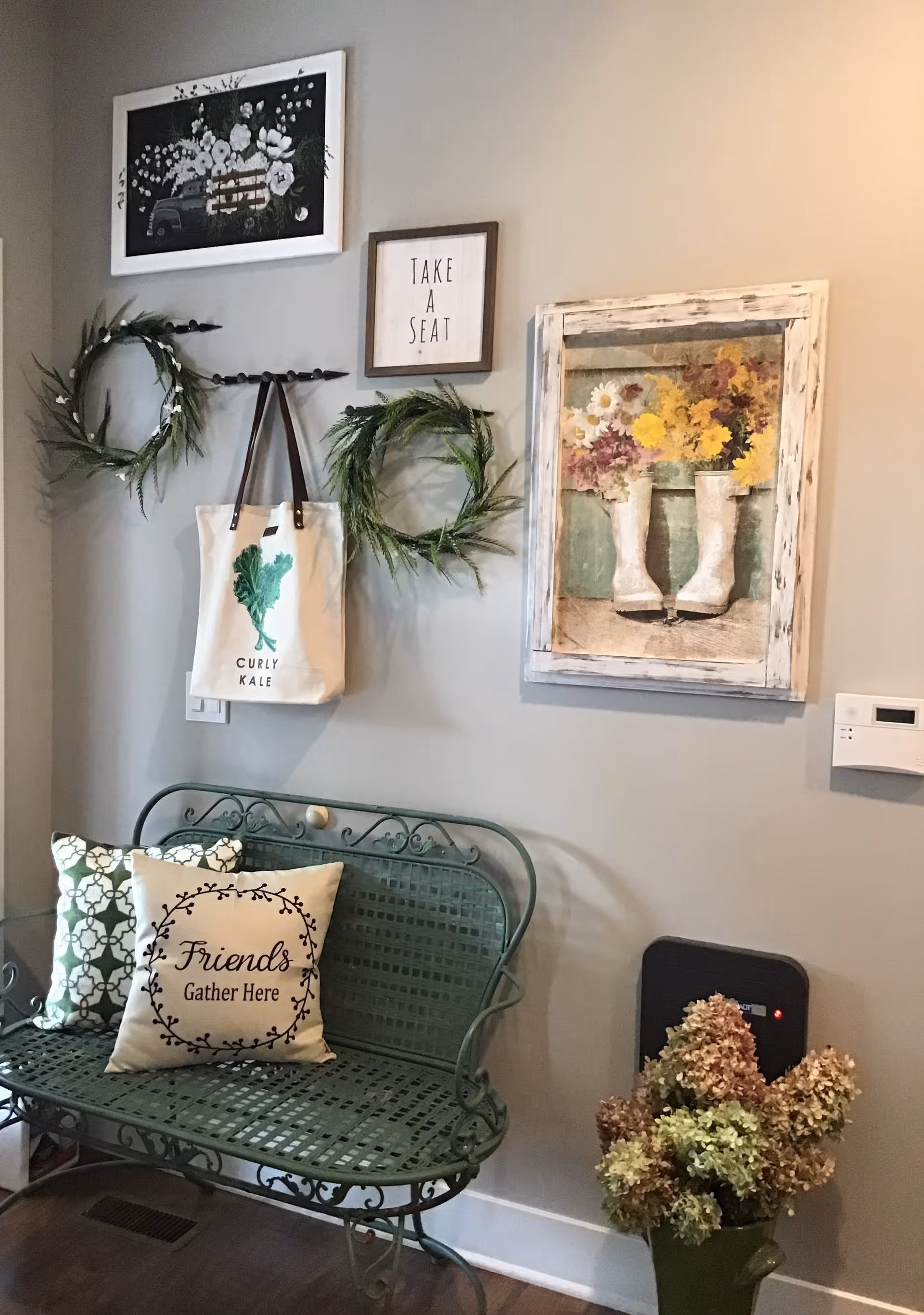 A decorative entry nook with a green metal bench with pillows, wreaths and framed art on the wall, and a vase of dried flowers on the floor.