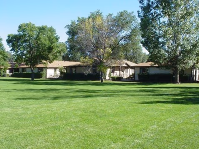 A senior living facility with a large, well-maintained green lawn in the foreground and several single-story buildings surrounded by mature trees in the background under a clear blue sky.