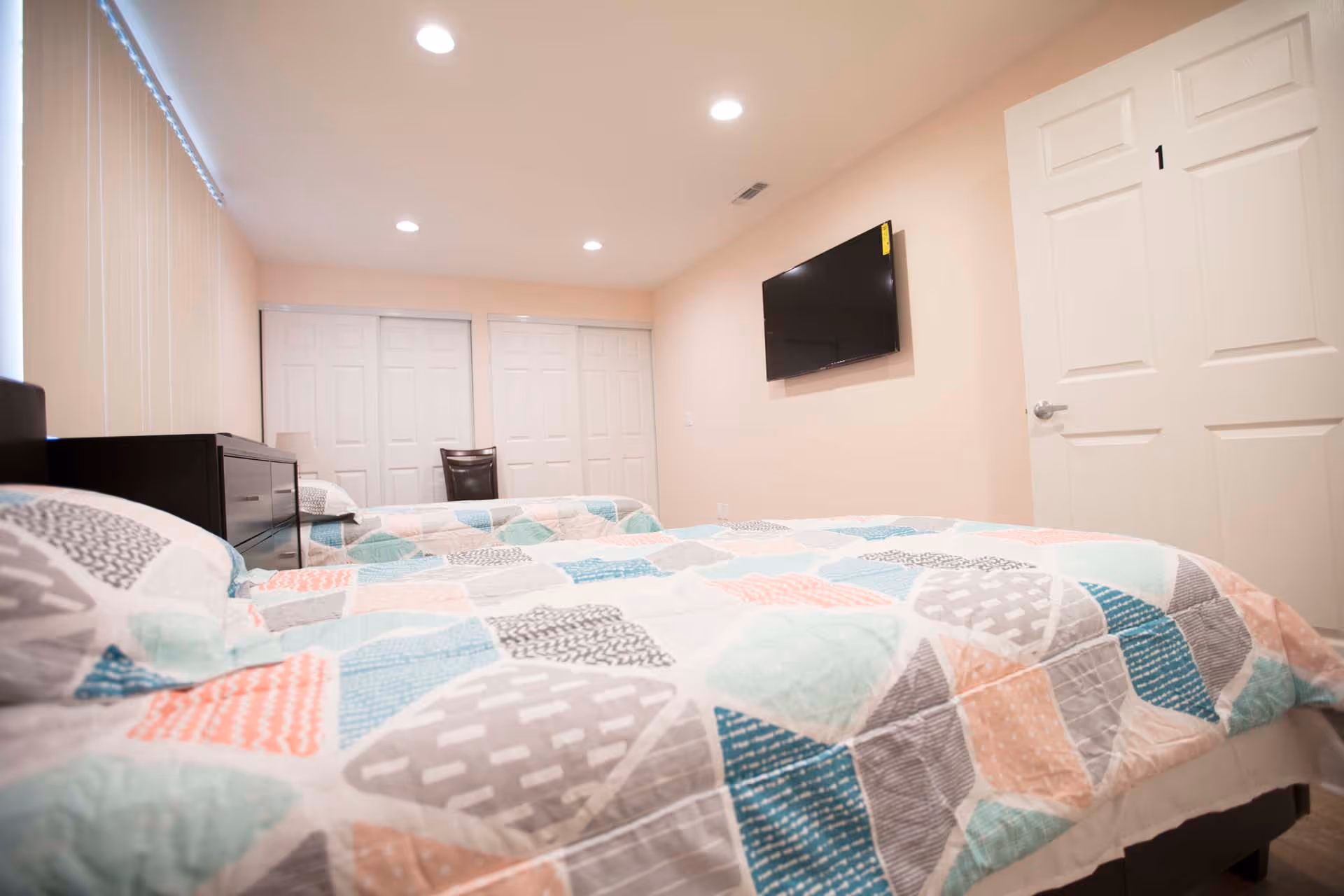Bright furnished bedroom with two beds covered in patterned quilts, a wall-mounted TV, dresser, and closet doors.