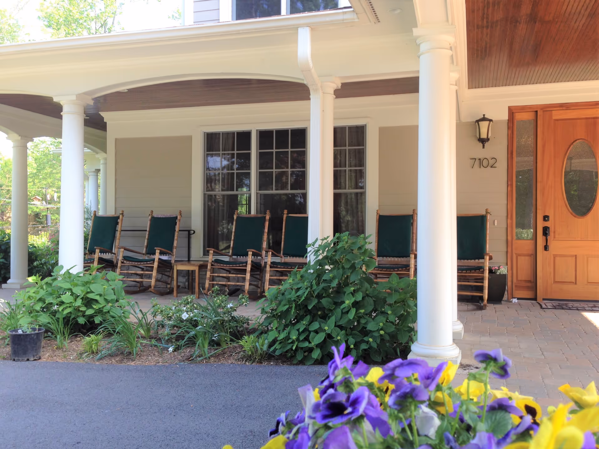 Front porch of a building with white columns, wooden rocking chairs with green cushions, a wooden door with an oval window, and a garden with green plants and colorful flowers in the foreground.
