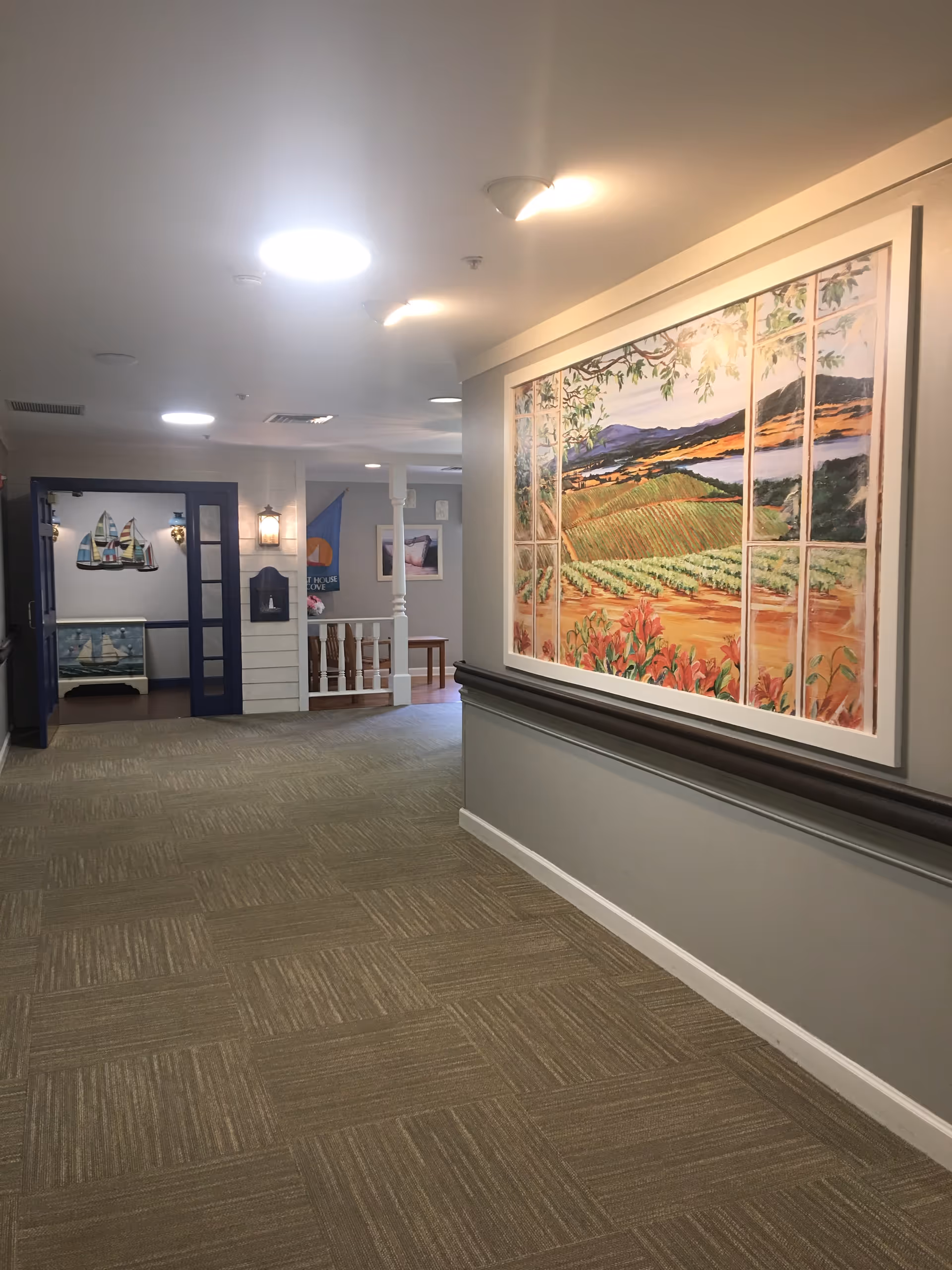 A well-lit hallway in an assisted living facility with beige carpet tiles and light gray walls. On the right wall, there is a large colorful painting depicting a scenic landscape with fields, hills, and a river. At the end of the hallway, there is an open doorway with blue double doors leading to another room decorated with nautical-themed artwork and furniture.