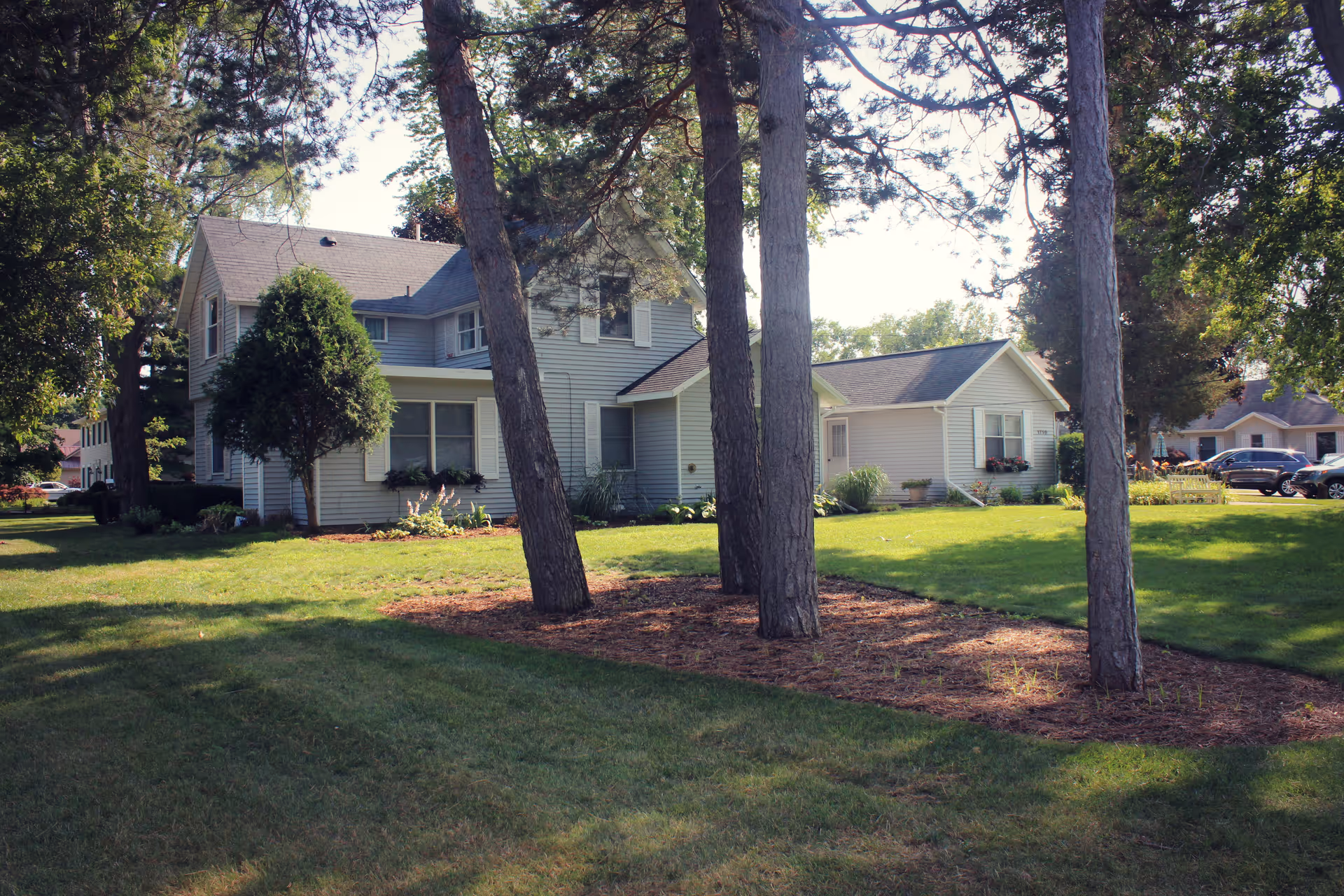 Two-story light-gray house with a manicured lawn, flowerbeds, and several tall trees in the front yard.