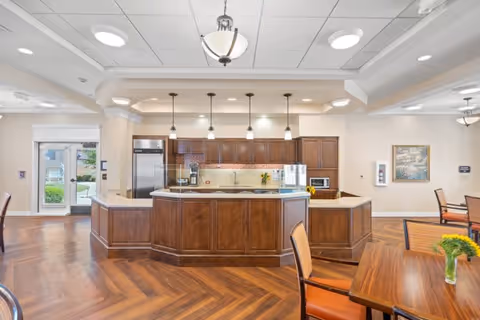 A spacious and well-lit kitchen area in a senior living facility with wooden cabinetry, a large island with a sink, stainless steel refrigerator, coffee machine, microwave, and pendant lighting. The room features herringbone wood flooring and adjacent dining tables with chairs. Large windows and glass doors provide natural light and a view of the outdoors.