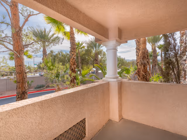 View from a covered balcony with beige stucco walls and a white column, overlooking palm trees and other greenery with a clear sky in the background.