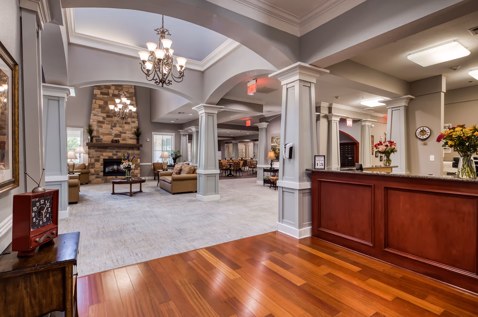 Large senior living lobby with a reception desk, seating area around a stone fireplace, columns, and chandeliers.