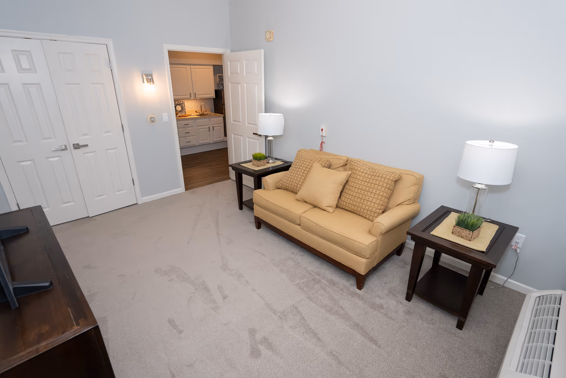A small living room with a beige couch adorned with three cushions, two dark wooden side tables each holding a lamp and a small plant, light gray walls, beige carpet, and a view into a kitchen area with white cabinets and wooden flooring.