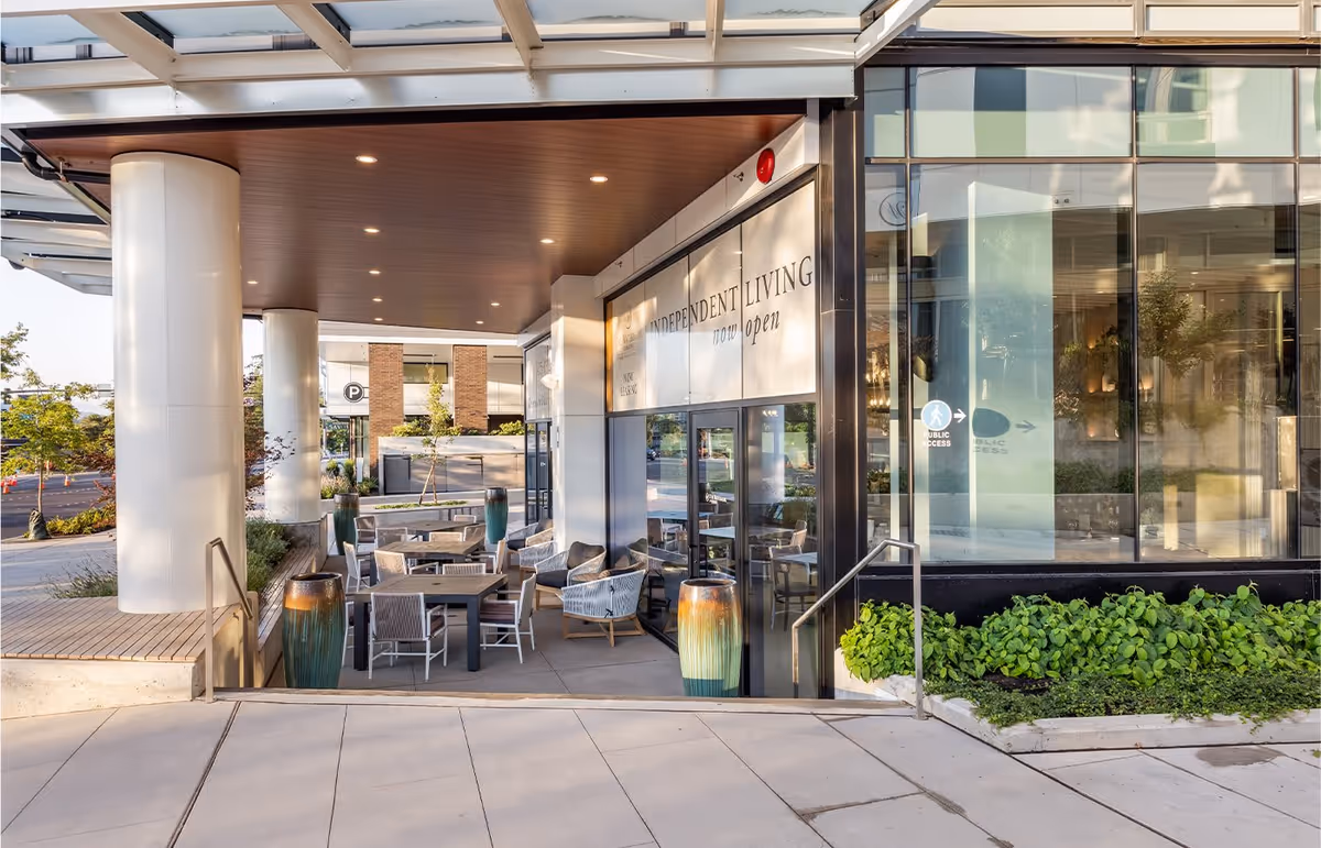 Outdoor covered seating area with tables and chairs next to a building entrance with large glass windows. Sign above the entrance reads 'Independent Living now open'. There are large white columns supporting the roof and decorative green plants along the walkway.