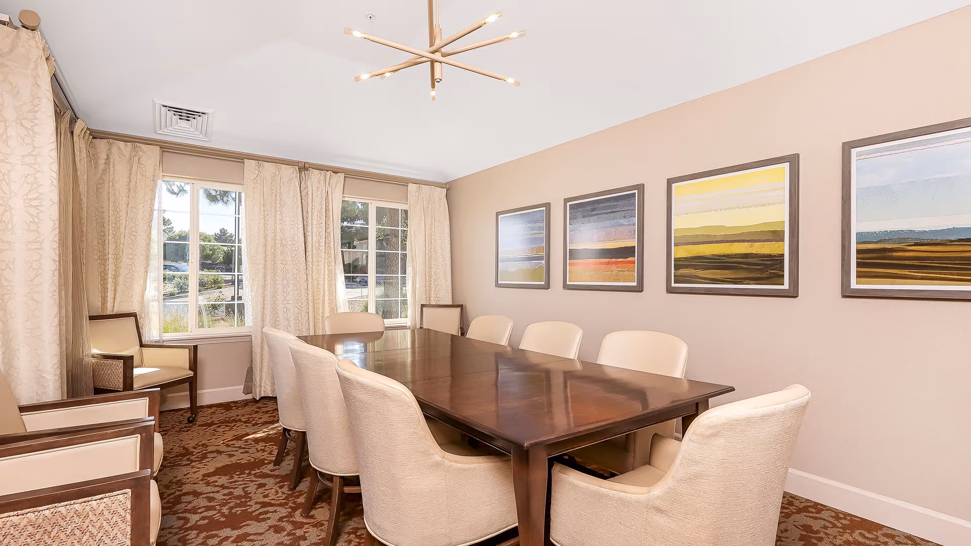 A well-lit dining room with a large rectangular wooden table surrounded by eight beige upholstered chairs. The room has beige walls adorned with four framed landscape paintings. Large windows with cream-colored curtains allow natural light to fill the space. There are additional chairs placed near the windows, and a modern chandelier hangs from the ceiling.