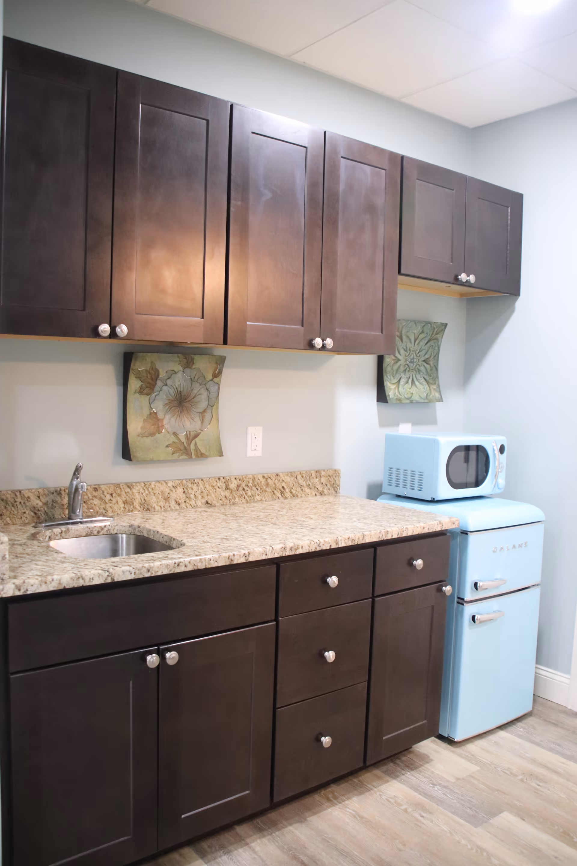 A small kitchen area with dark brown cabinets, a granite countertop, a stainless steel sink, a light blue retro-style mini refrigerator, and a matching light blue microwave on top. The walls are light blue with two floral decorative tiles mounted above the countertop.