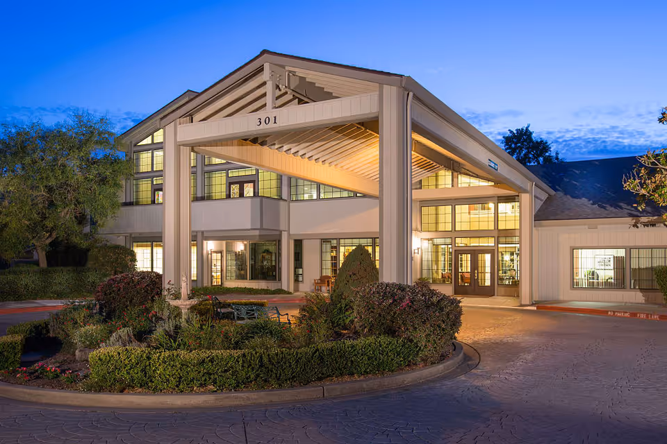 Exterior view of Oakmont Gardens facility at dusk, showing a well-lit entrance with large windows, a covered drop-off area, and landscaped greenery in front.