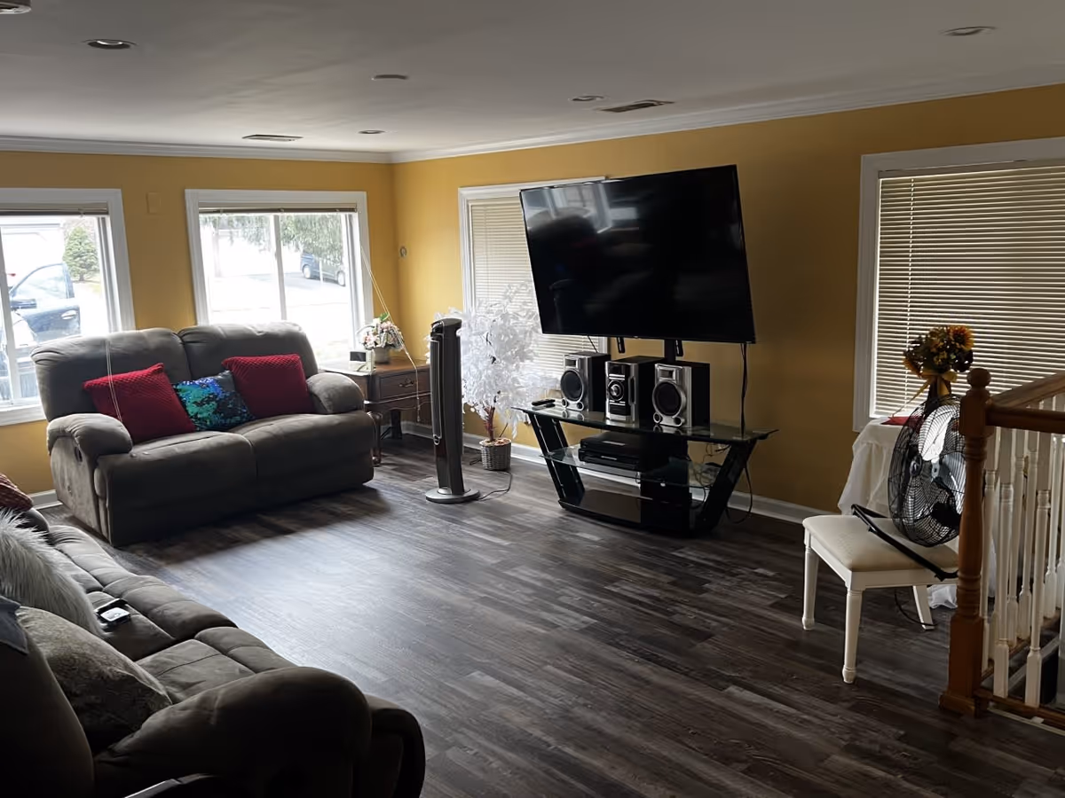 A living room with two gray sofas, one with red and blue pillows, large windows with white blinds, a flat-screen TV mounted on a black glass stand with a sound system, a standing fan on a white chair, a small wooden side table with a plant, and yellow painted walls.