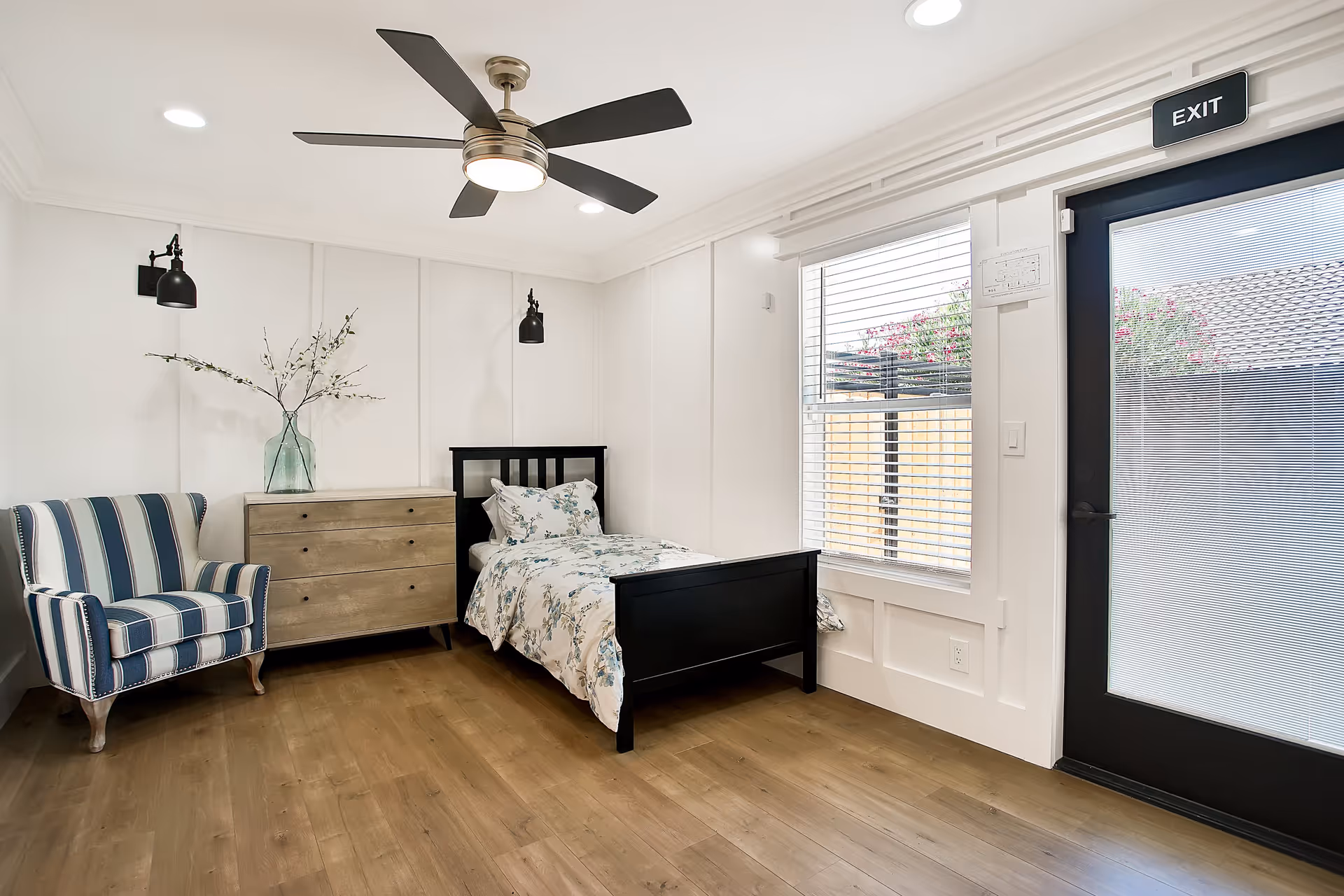Bright, modern bedroom with a single black bed, striped armchair, wooden dresser, ceiling fan, and an exit door with window.