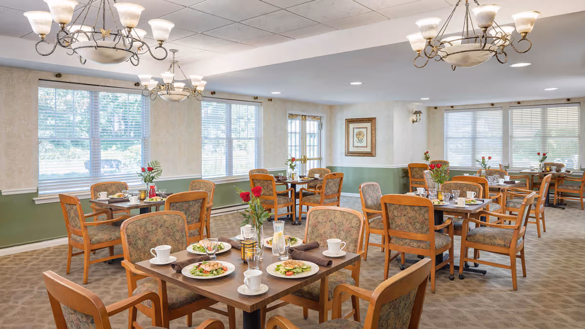 A bright and spacious dining room with multiple tables set for meals. Each table has plates with food, cups, glasses, and floral centerpieces. The room features large windows with blinds, green and beige walls, carpeted floors, and elegant chandeliers hanging from the ceiling.