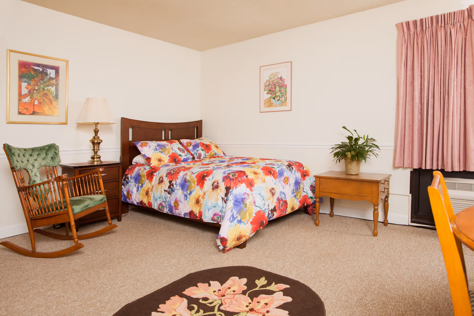 A cozy bedroom with a bed featuring a colorful floral bedspread and matching pillows. To the left of the bed is a wooden nightstand with a brass lamp and a green cushioned rocking chair. On the right side, there is a wooden side table with a potted plant. The room has beige carpet, white walls with two framed floral paintings, and pink curtains covering a window.