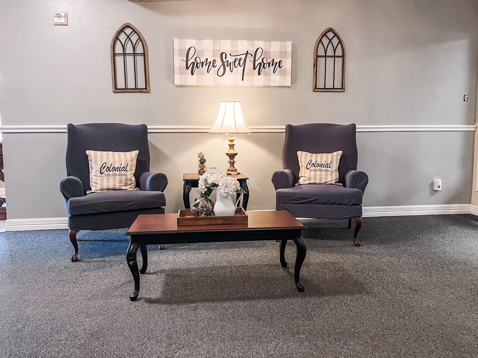 A cozy sitting area with two dark upholstered armchairs, each with a beige and white striped pillow that reads 'Colonial Nursing & Rehabilitation.' Between the chairs is a small wooden side table with a decorative lamp and ornaments. In front of the chairs is a wooden coffee table with a white vase of flowers and decorative spheres. On the wall behind the chairs is a sign that says 'home sweet home' flanked by two decorative window frame wall hangings.