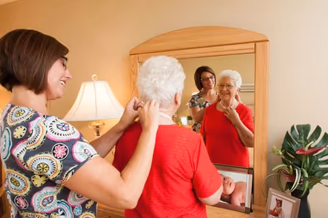 A woman is helping an elderly woman put on a necklace in front of a mirror in a warmly lit room with a lamp and a plant on a dresser.