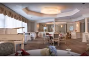 A spacious and elegantly decorated dining room with round tables covered in white tablecloths, each set with glassware and napkins. The room features a grand piano on the left side, large windows with draped curtains allowing natural light, and a chandelier hanging from a tray ceiling. The walls are painted in soft neutral tones with decorative moldings.