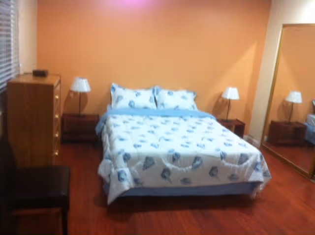 A bedroom with a double bed covered in a white and blue seashell patterned bedspread. There are two matching pillows on the bed. On either side of the bed are wooden nightstands with white lamps. To the left of the bed is a wooden dresser with several drawers and a small black item on top. A black chair is positioned near the dresser. The room has wooden flooring and peach-colored walls. A large mirror is mounted on the right wall, reflecting part of the bed and lamp.