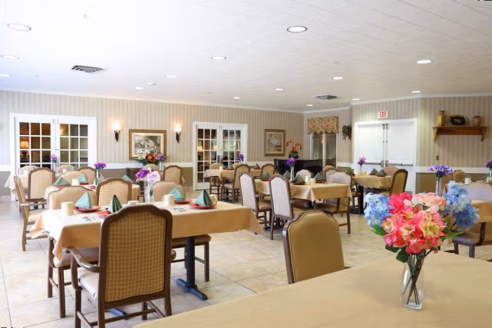 A bright and spacious dining room with multiple tables covered in beige tablecloths, each set with folded green napkins, plates, and cups. The room features cushioned chairs with wooden frames, floral centerpieces in vases on each table, beige tiled flooring, and soft lighting from wall sconces and ceiling lights. French doors and framed paintings adorn the walls.