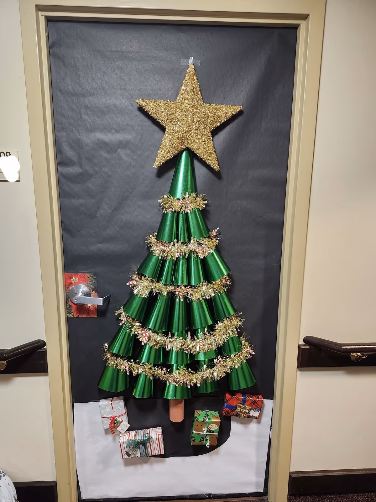 Interior door decorated like a Christmas tree with layered green cones, a gold star topper, tinsel garlands and wrapped presents at the base.
