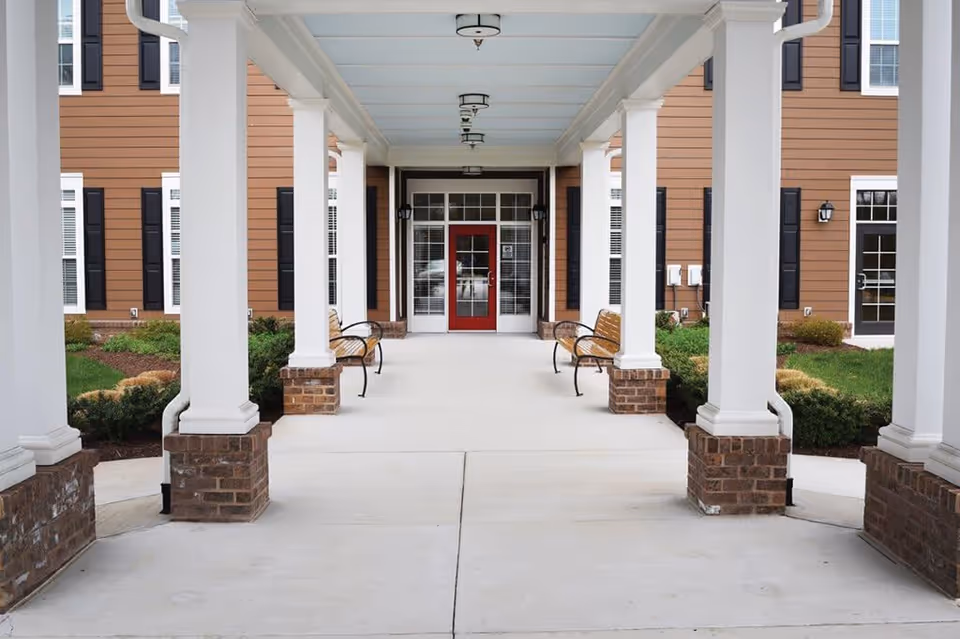 Covered entrance walkway with white columns and brick bases leading to a building with a red door and windows. There are wooden benches with black metal armrests on either side of the walkway, and landscaping with green shrubs and grass around the building.