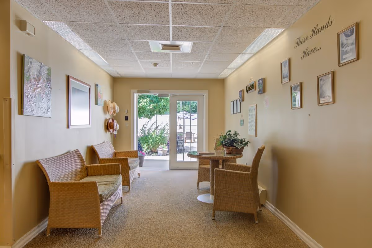 A cozy indoor seating area with wicker chairs and a small round table with a potted plant. The walls are decorated with framed pictures and hats hanging on hooks. A glass door at the end of the room opens to an outdoor patio with greenery.