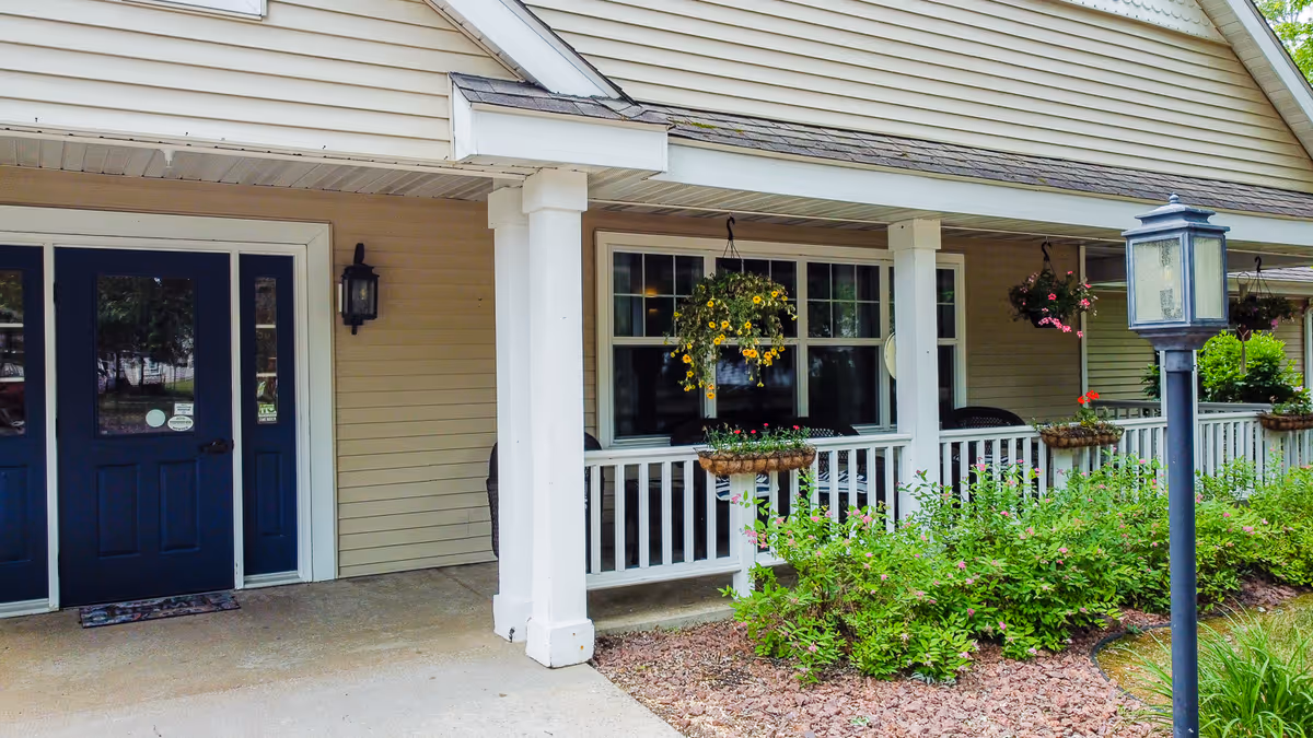 Exterior view of a senior living facility entrance with beige siding, a covered porch supported by white columns, hanging flower baskets, and a black lamp post in the landscaped garden area.