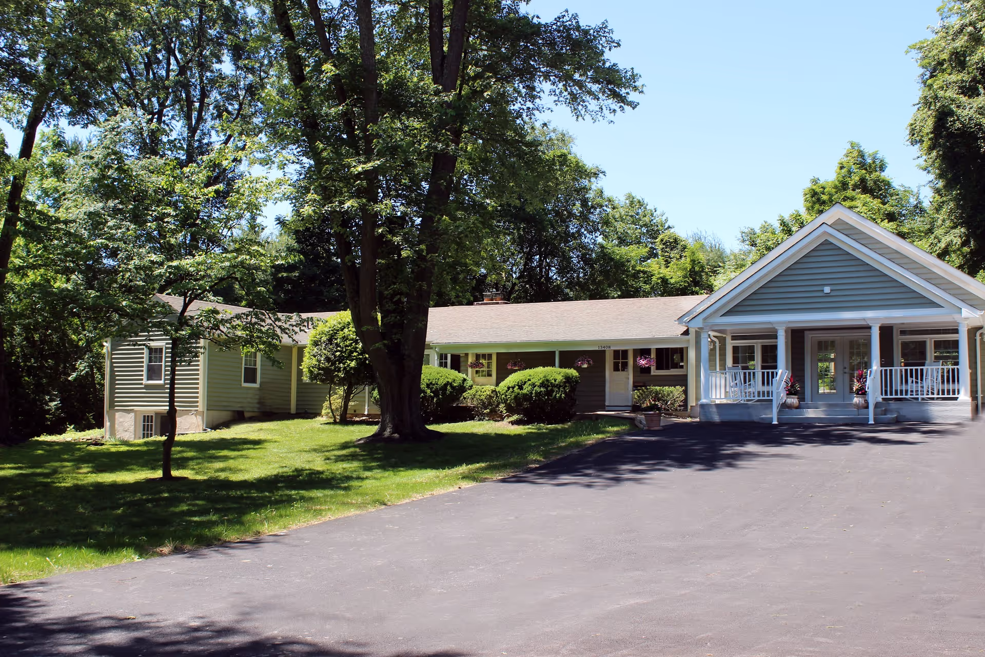 Exterior view of a single-story residential building with a covered porch and white railings, surrounded by green trees and bushes under a clear blue sky.