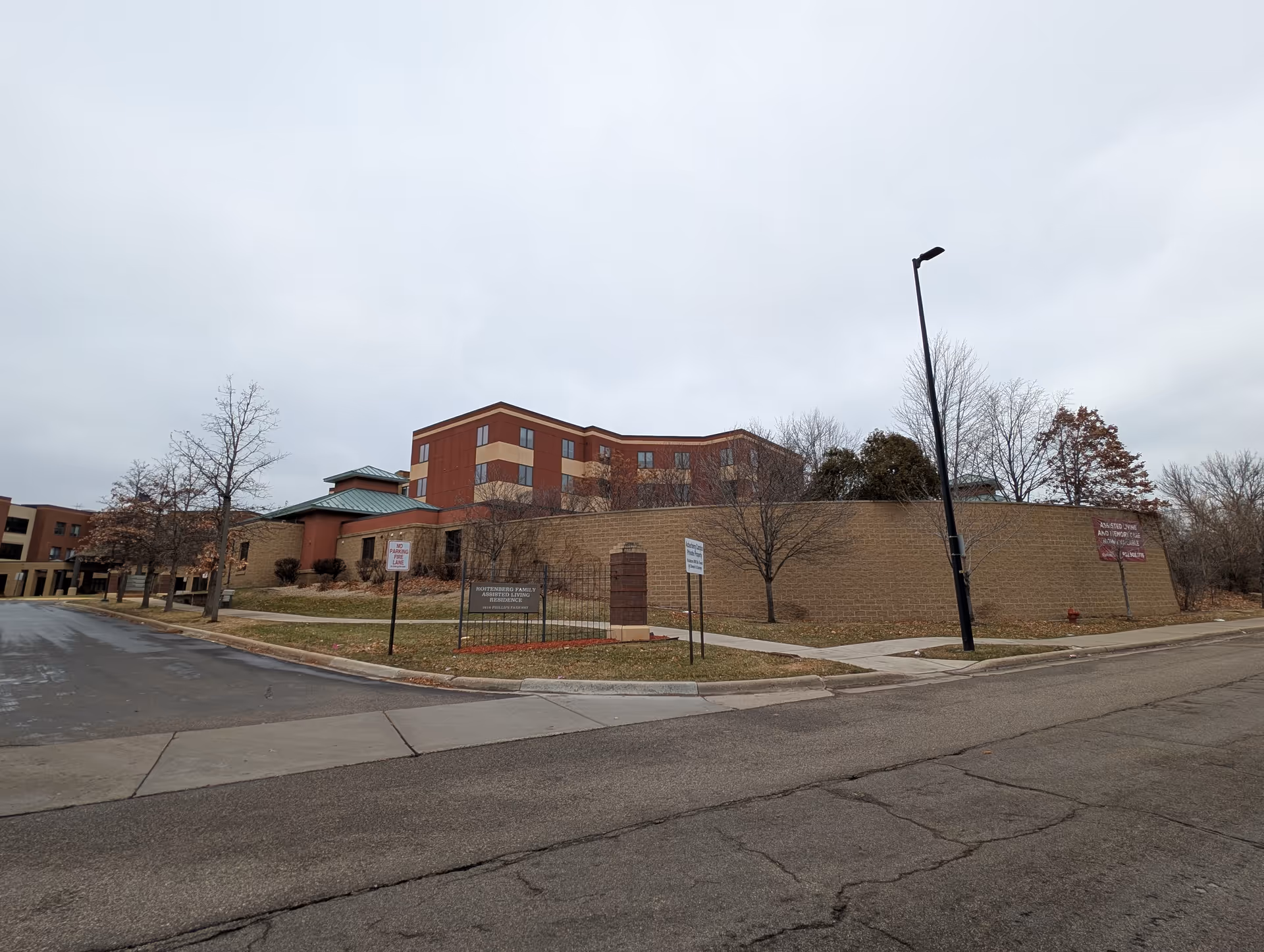 Exterior view of Roitenberg Family Assisted Living building with a multi-story structure behind a brick wall, leafless trees, a street lamp, and a road in the foreground under a cloudy sky.